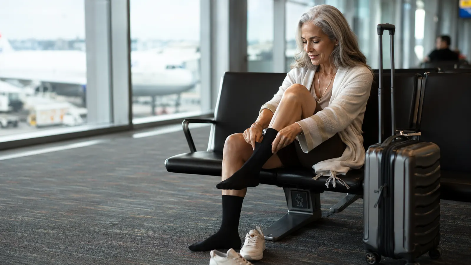 Woman in her sixties sitting in an airport gate seat, rolling merino compression socks onto her feet before boarding, carry-on bag beside her