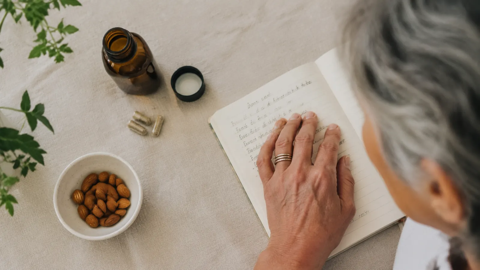 Overhead flat lay of an amber supplement bottle, an open sleep journal with handwritten notes, and a small bowl of almonds on cream linen cloth, warm diffuse natural light